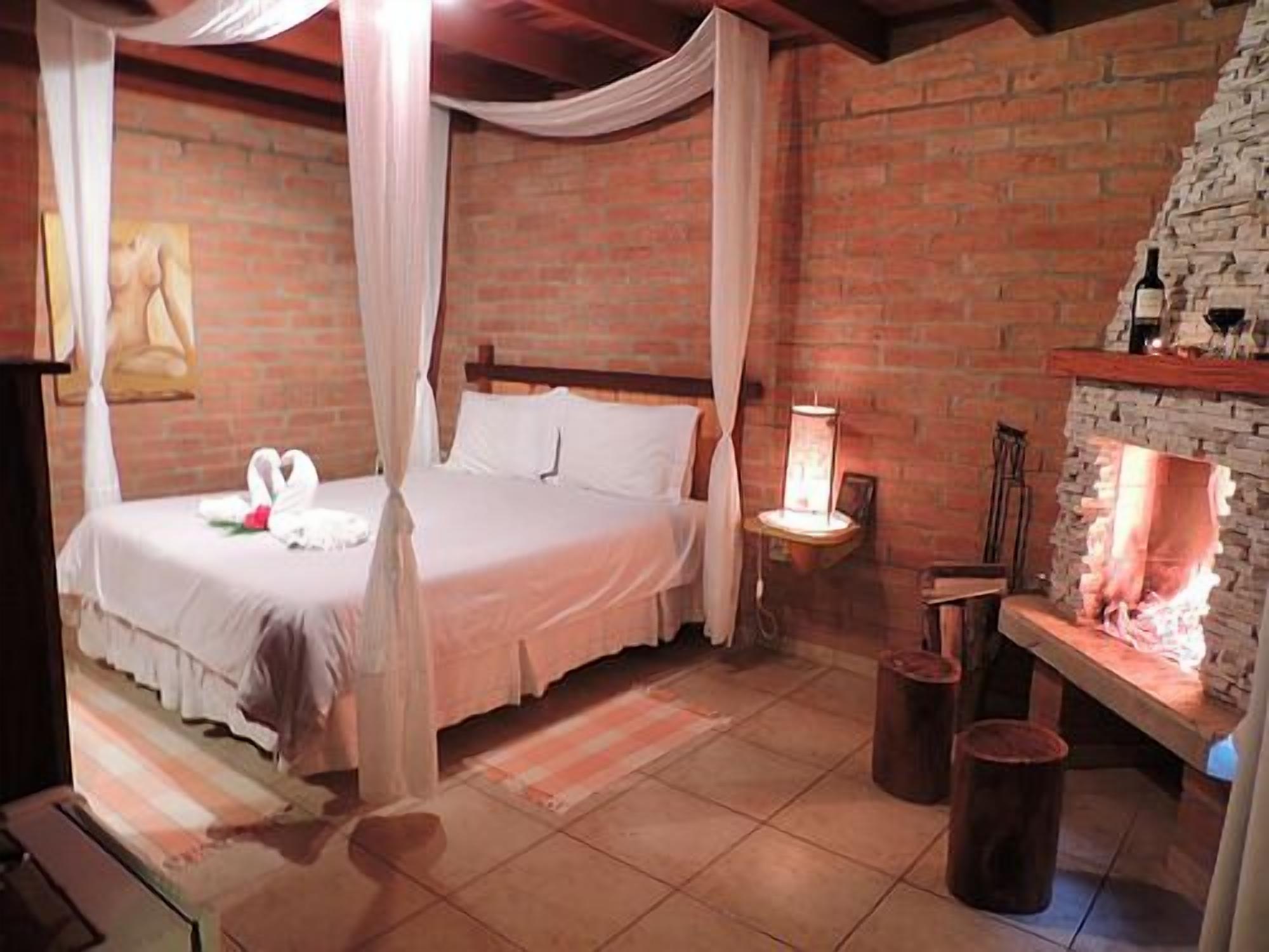 Un dormitorio con pared de ladrillo, cama con ropa de cama blanca, mesita de noche con lámpara y chimenea.