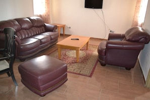 A living room with a brown leather sofa, a matching armchair, a wooden coffee table, and a television mounted on the wall. - Meumi Residence (Yaoundé)