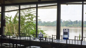 A dining area with tables set for two, overlooking a river with dense foliage and trees in the background. - Amazon Planet (Tambopata)