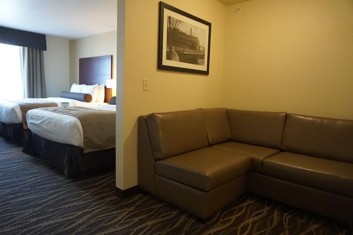 Ein Hotelzimmer mit einem großen braunen Ledersofa, zwei Betten mit weißen Bettwäsche und einem gerahmten Bild an der Wand, sowie einem gemusterten Teppich.