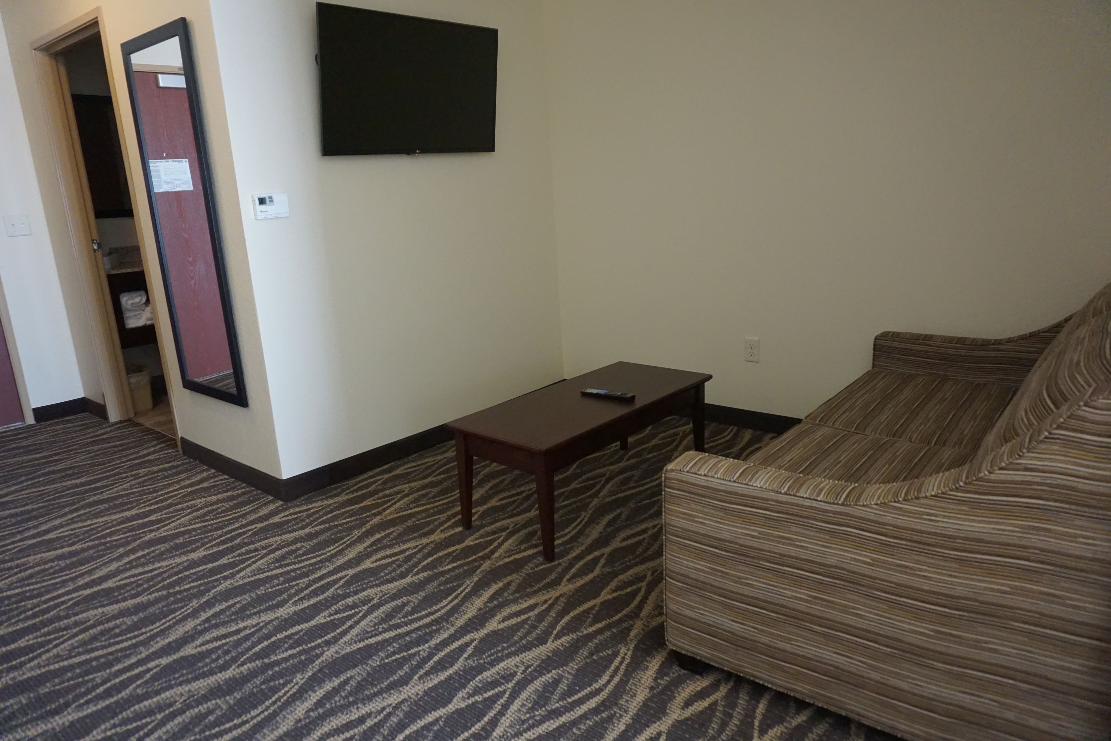 A hotel room with a striped sofa, a small wooden table, a flat-screen TV mounted on the wall, and a carpeted floor.