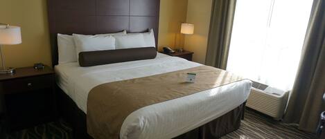 Una habitación de hotel con una cama grande, dos mesitas de noche, un armario auxiliar, un aire acondicionado instalado en la pared y una ventana con cortinas.
