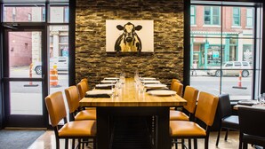 Un restaurant avec une longue table en bois dressée pour un repas, un mur en pierre et un tableau représentant une vache accroché au mur.