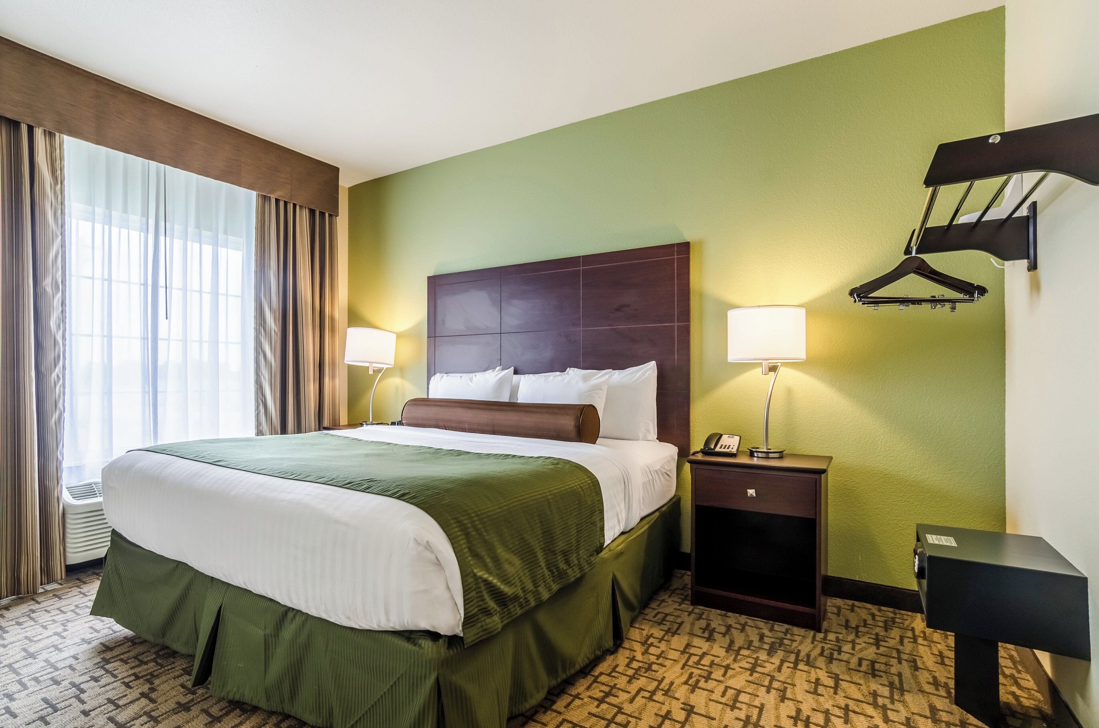 A hotel room with a large bed, two bedside lamps, a nightstand, a coat rack, and a window with curtains.