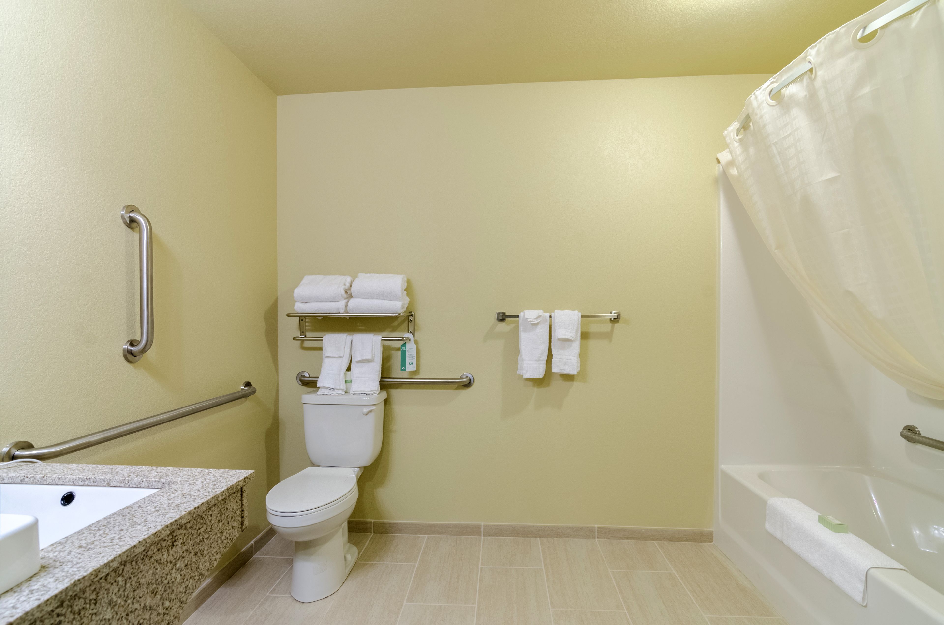 Un baño con inodoro, lavamanos y bañera. Hay barras de apoyo, toallas y una cortina de ducha.