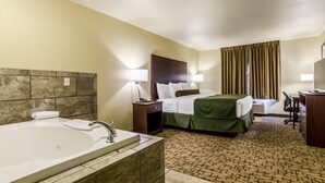 A hotel room with a large bathtub, a bed with a green bedspread, a desk with a chair, and a window with curtains.