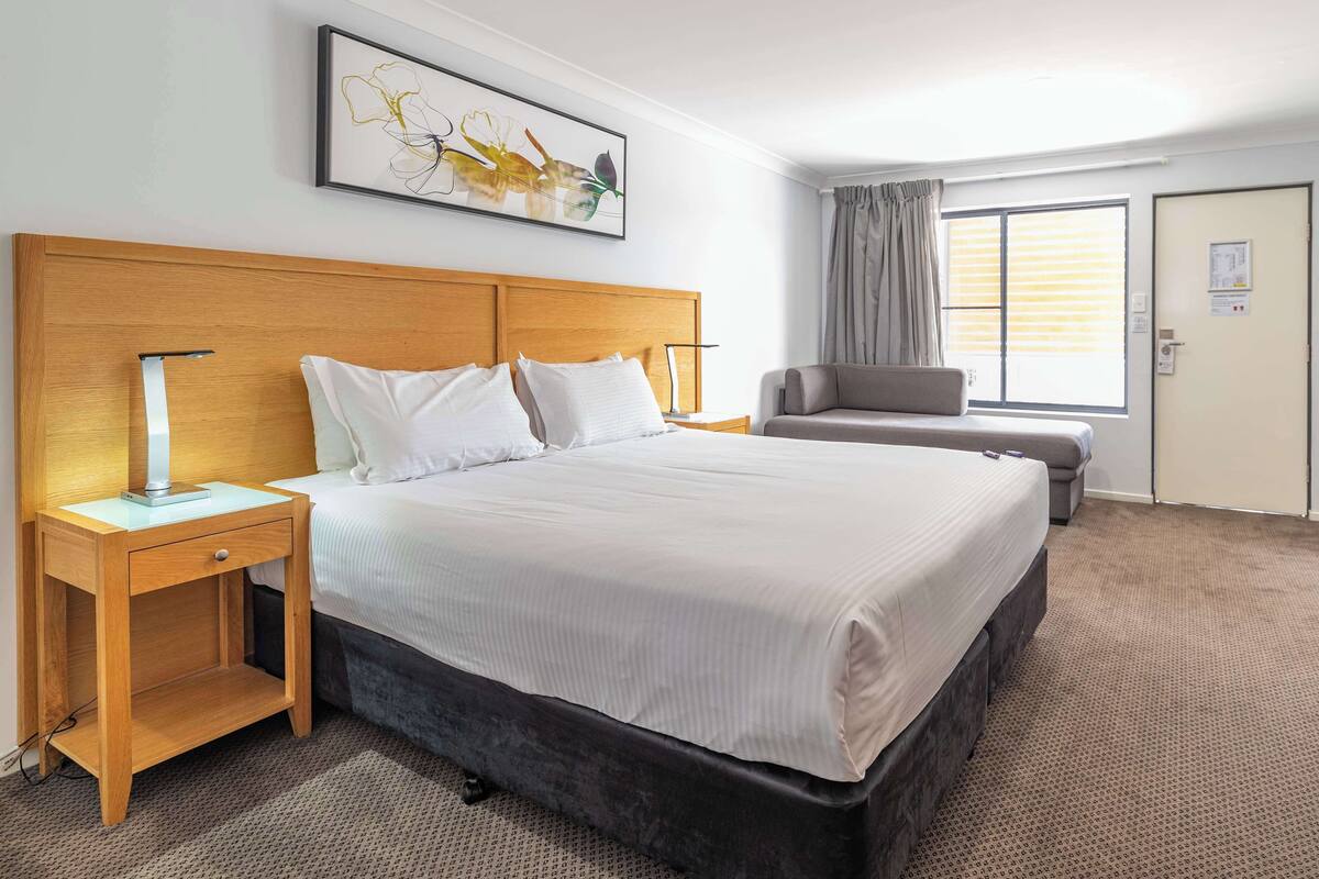 Ein Hotelzimmer mit einem großen Bett, einem Nachttisch aus Holz, einem Schreibtisch, einem Stuhl, einem Fenster mit Vorhängen und einem gerahmten Bild an der Wand.