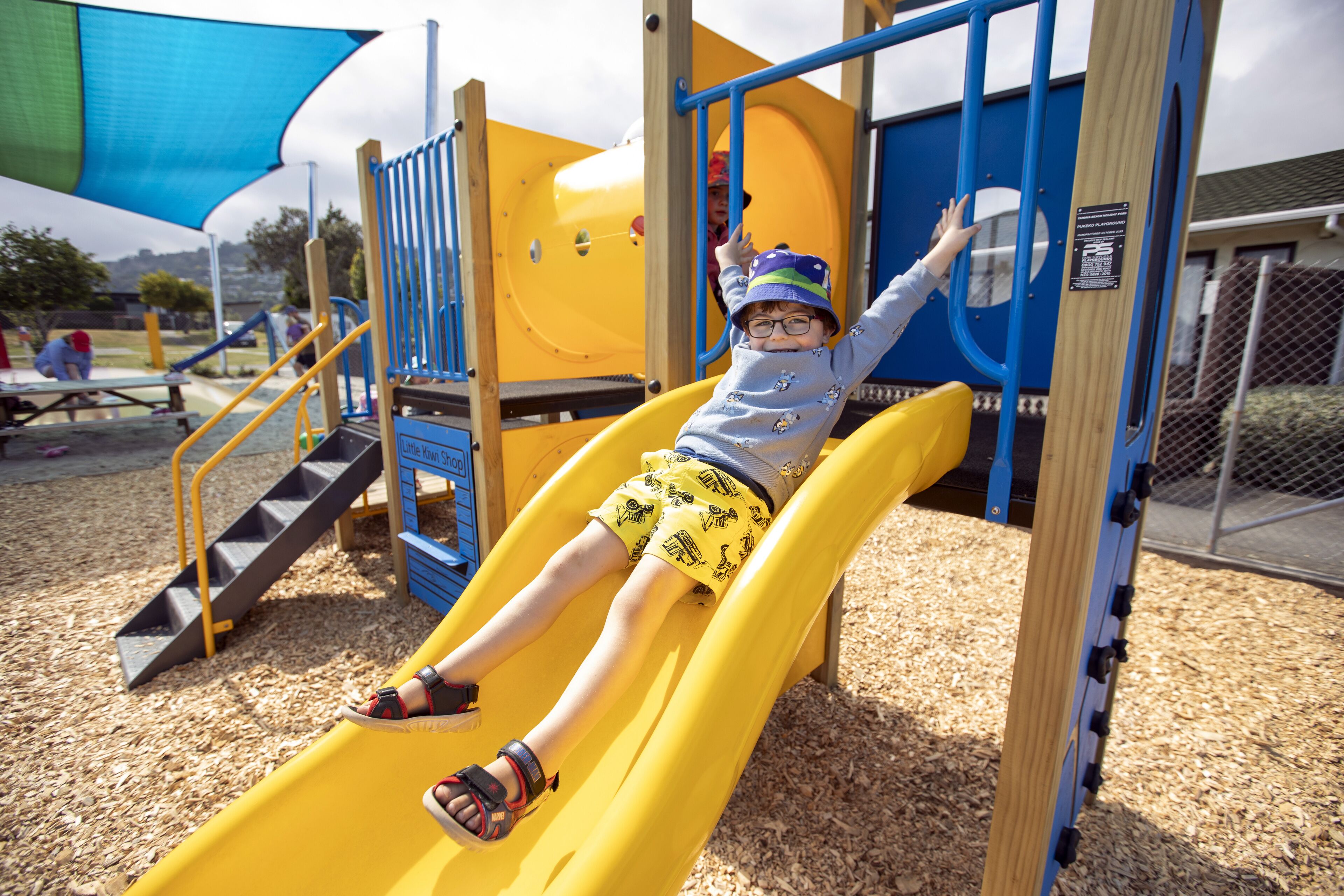 A child on a yellow slide at a playground with wooden structures and a blue canopy.
