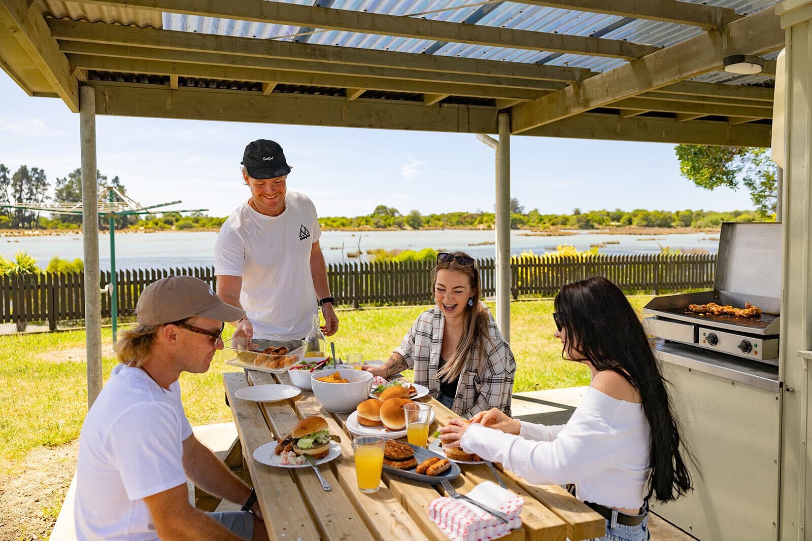A group of people gathered around a picnic table under a covered area with a grill.