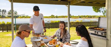 A group of people gathered around a picnic table under a covered area with a grill.