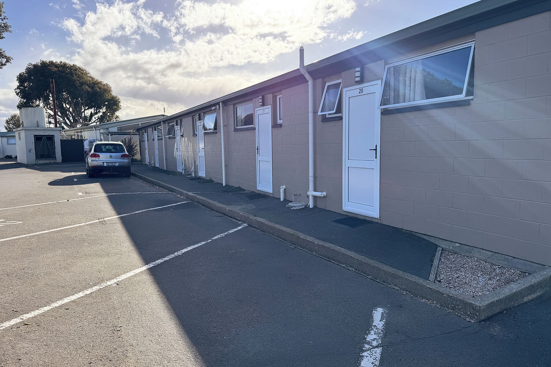 Een rij motelkamers op een verdieping, elk met een eigen deur, een parkeerplaats en een boom op de achtergrond.
