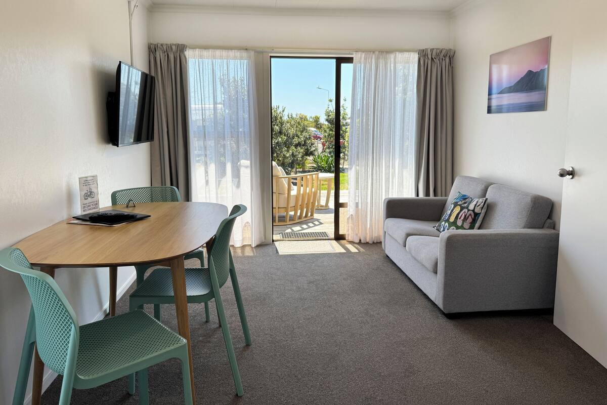 A room with a wooden table, green chairs, a grey sofa, and a TV mounted on the wall.