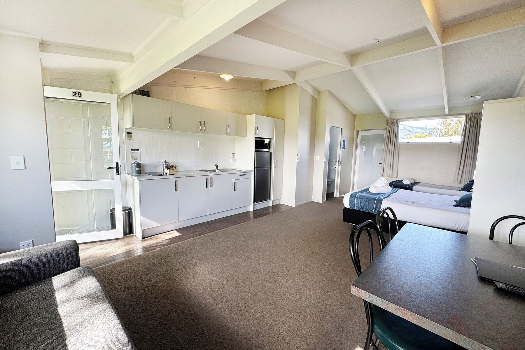 A compact kitchen with white cabinets, a refrigerator, and a sink. There is a dining area with a table and chairs, and a bedroom with a bed and window.