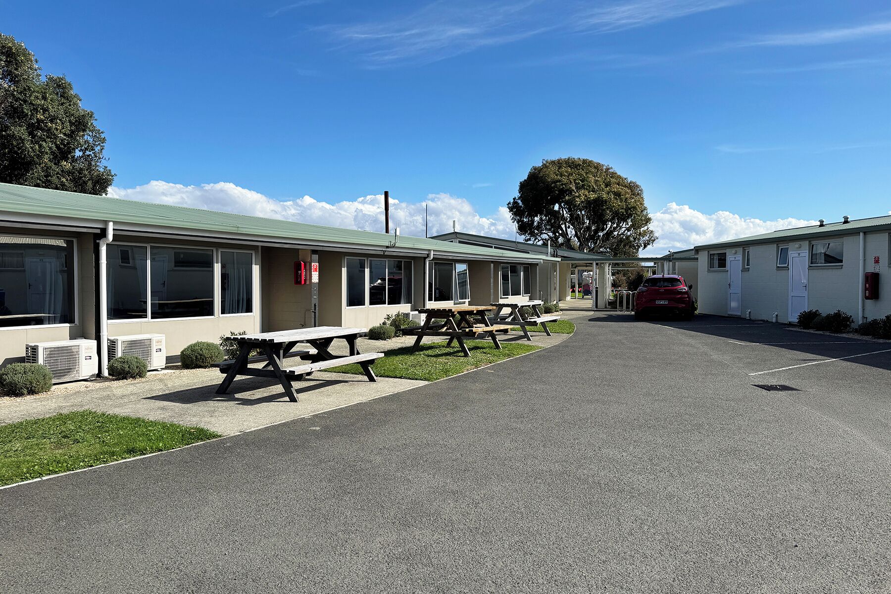 A row of single-story mobile homes with picnic tables in front, a paved driveway, and a red car parked in the lot.