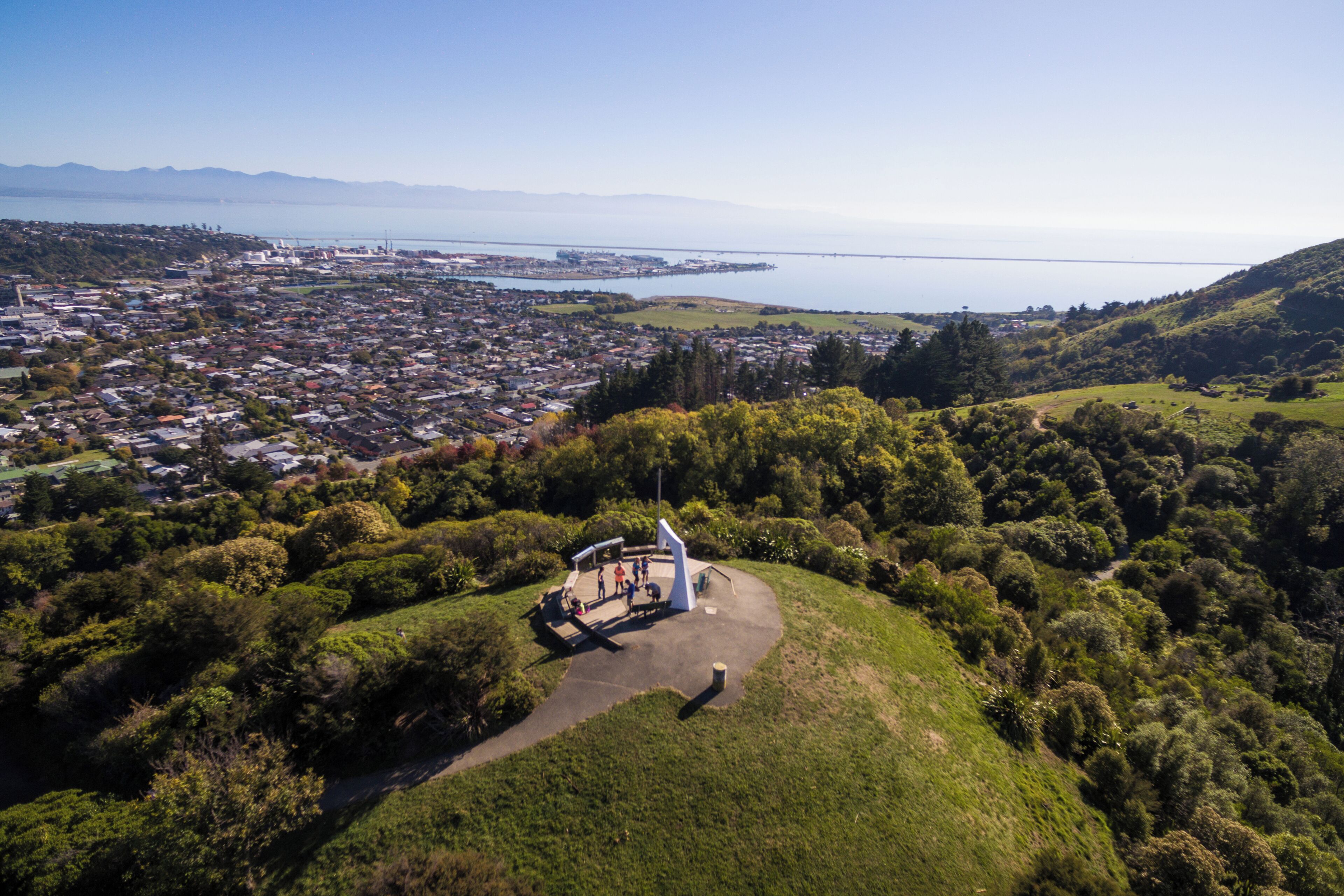 An aerial view of a coastal town with a helipad on a hill, surrounded by greenery and overlooking a bay.