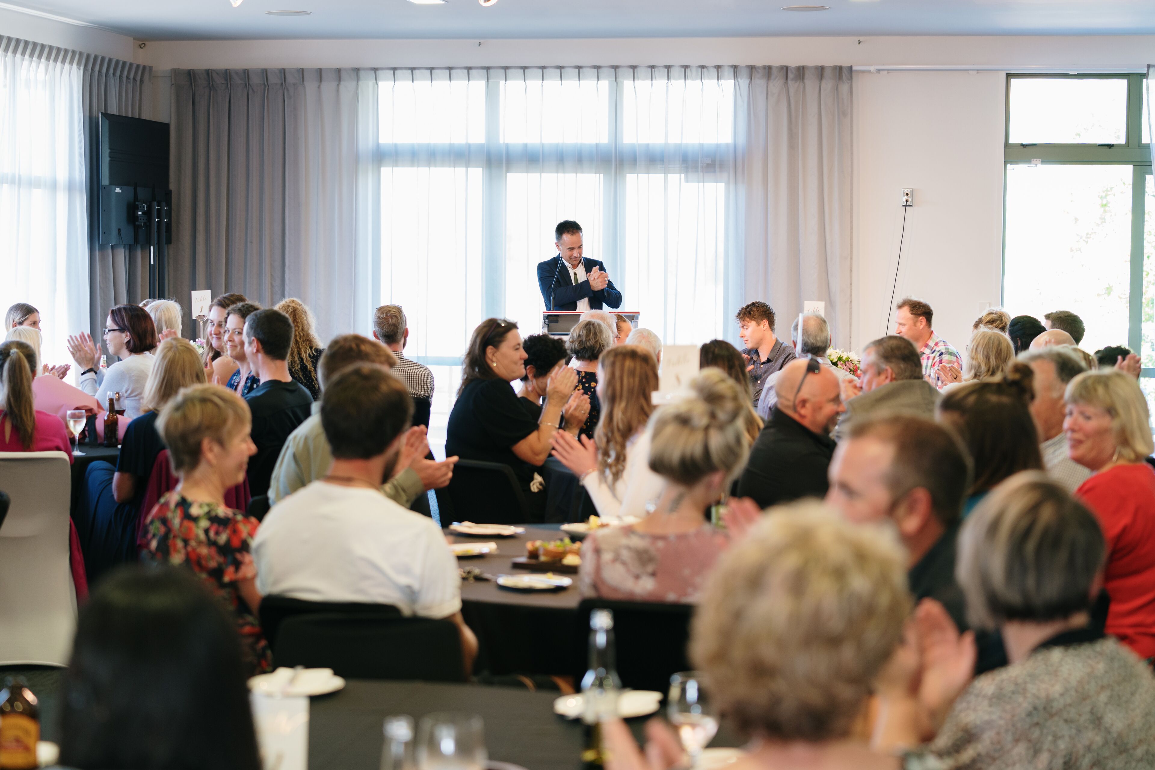 A conference room with a speaker at a podium, attendees seated at tables, and large windows with curtains.
