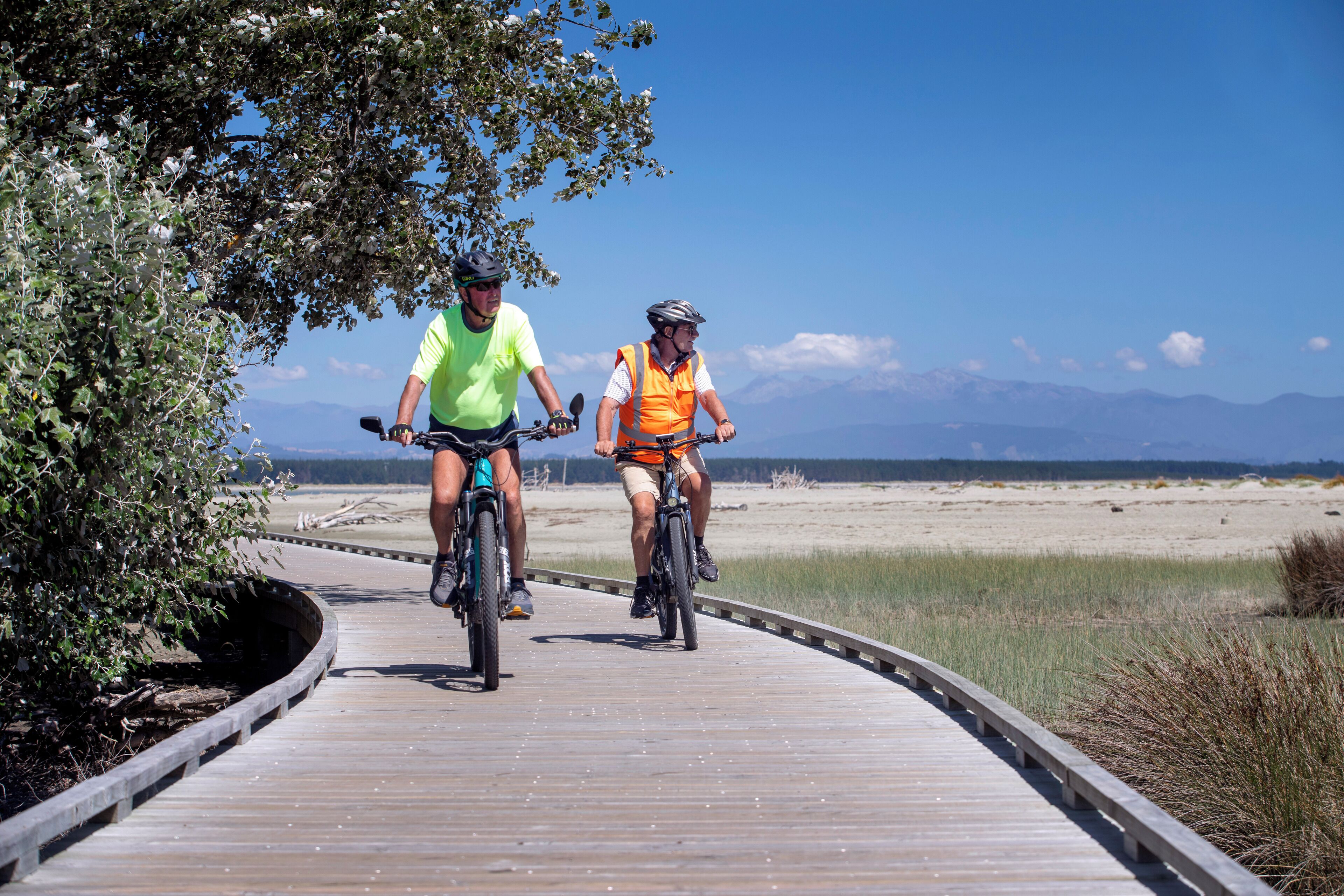 Two cyclists on a wooden boardwalk with a beach and mountains in the background.