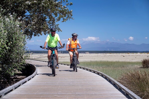 Dos ciclistes a dalt d'un camí de fusta davant d'una platja i muntanyes al fons.