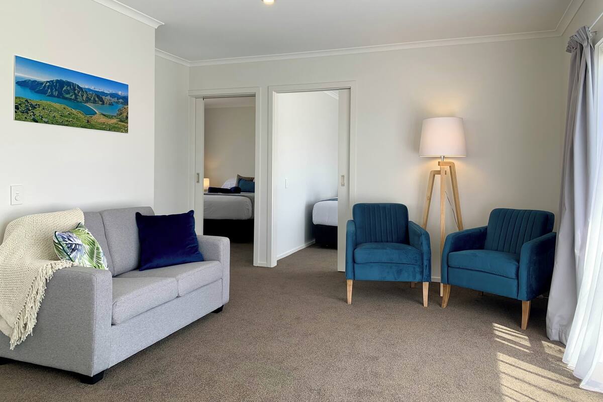 A hotel room with a grey sofa, two blue armchairs, a floor lamp, and a bed in the background.