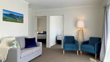 A hotel room with a grey sofa, two blue armchairs, a floor lamp, and a bed in the background.