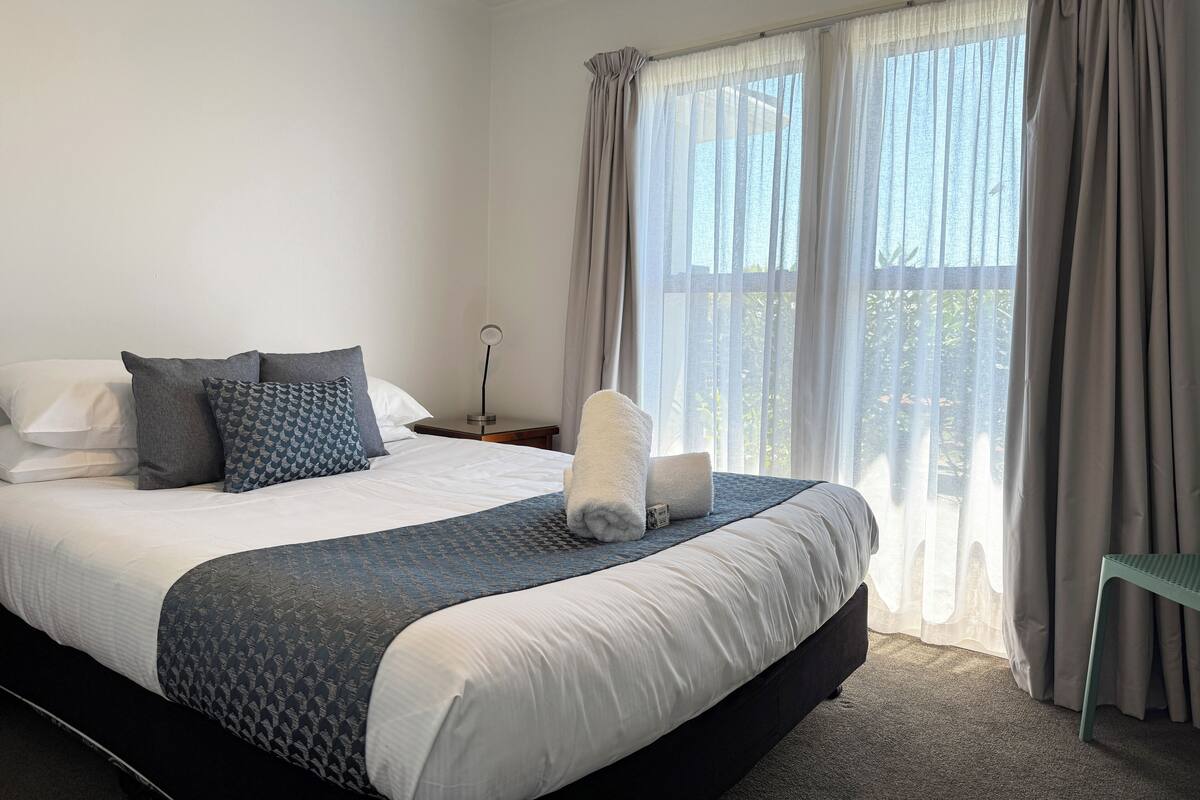 A neatly made bed with a white and dark blue patterned bedspread, a bedside lamp, and a view of the outdoors through sheer curtains.