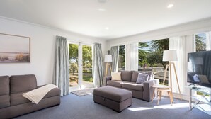 A modern living room with a grey sofa, a grey ottoman, and a glass coffee table. There is a painting on the wall and a window with curtains. - Tahuna Beach Holiday Park  (Nelson)