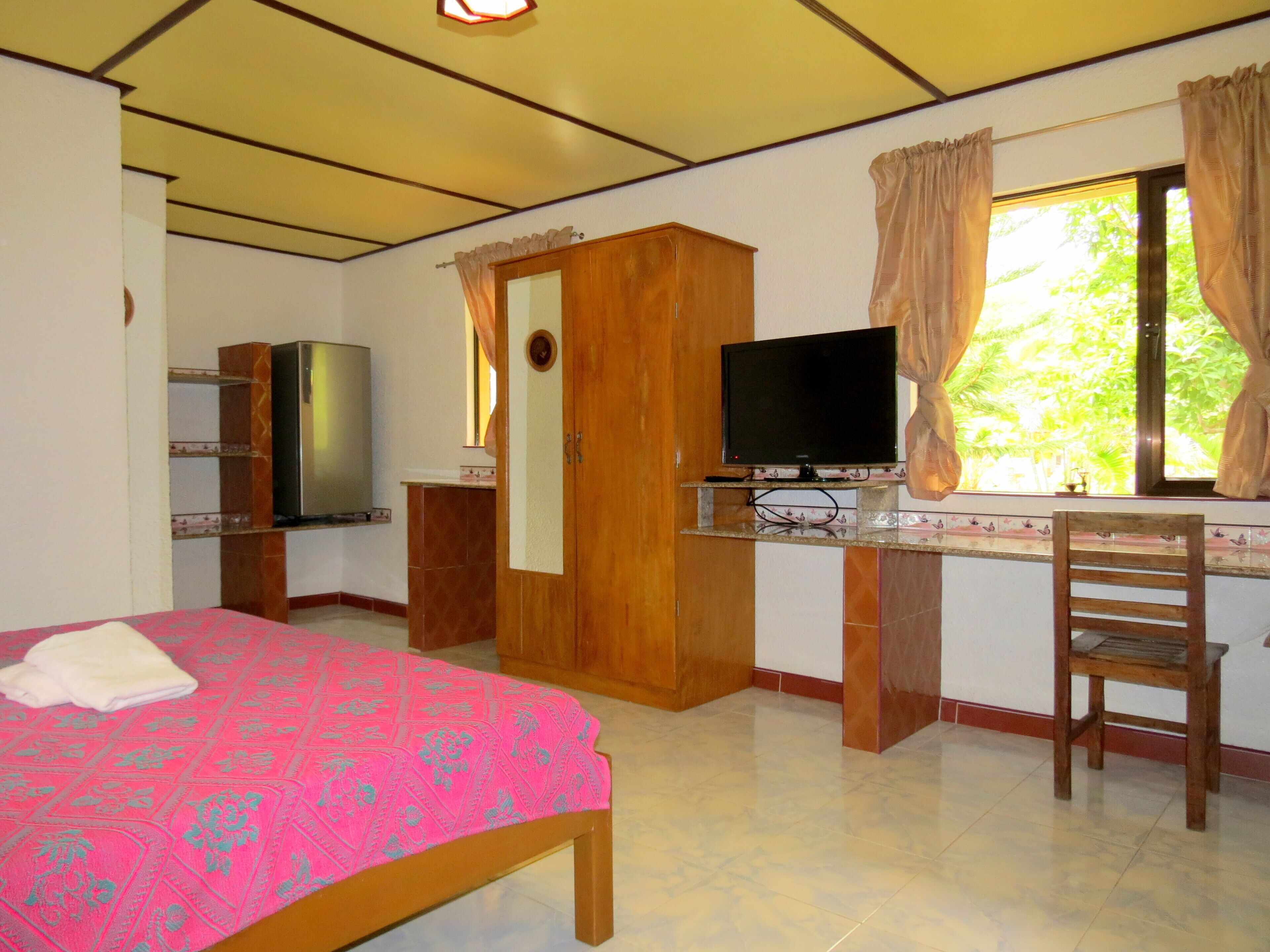 A bedroom with a bed, a TV, a wooden wardrobe, and a window with curtains.