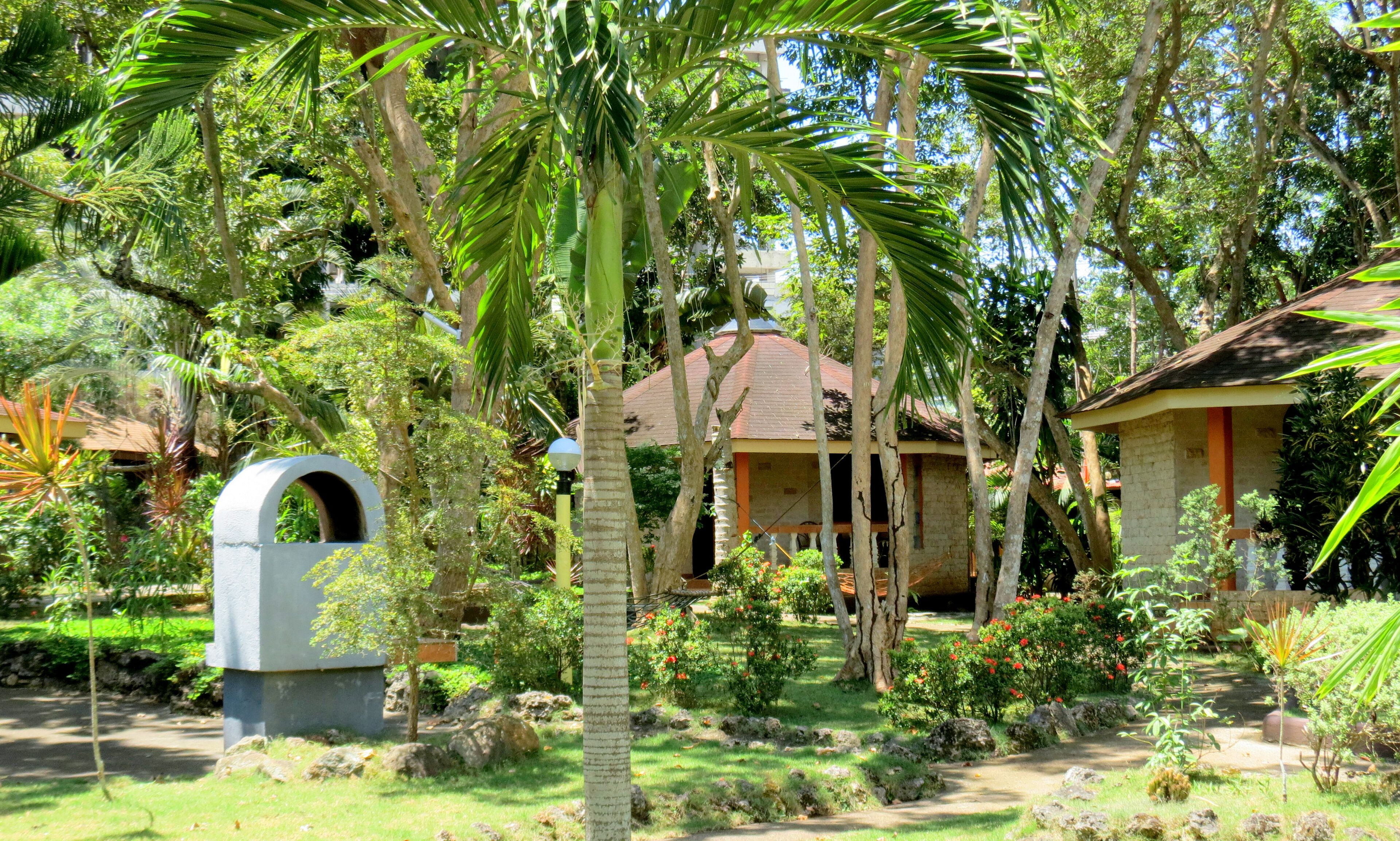Une maison traditionnelle avec un toit en tuiles, entourée d’une végétation luxuriante et de palmiers.