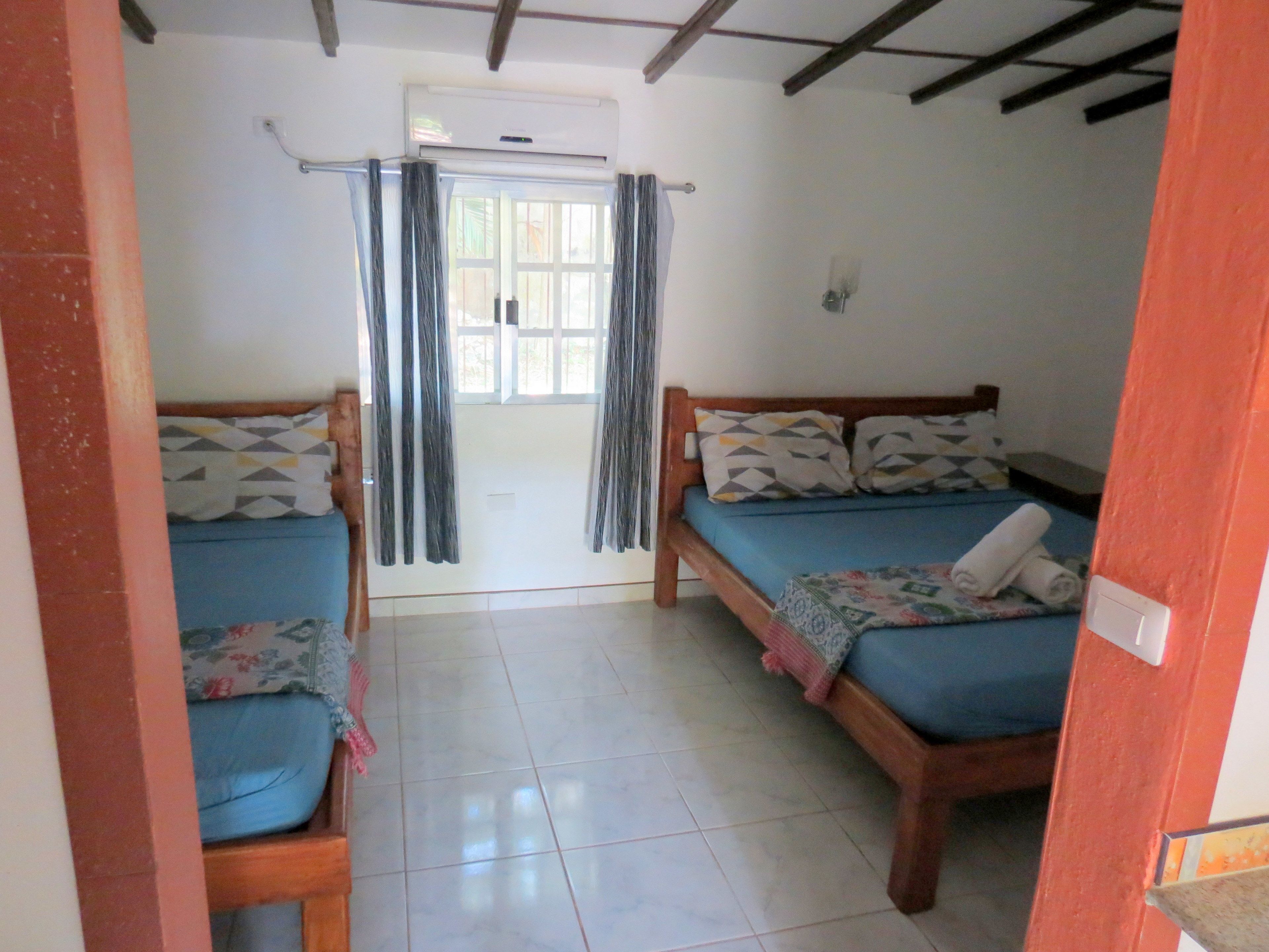 A room with two wooden beds, a tiled floor, a window with curtains, and an air conditioning unit.