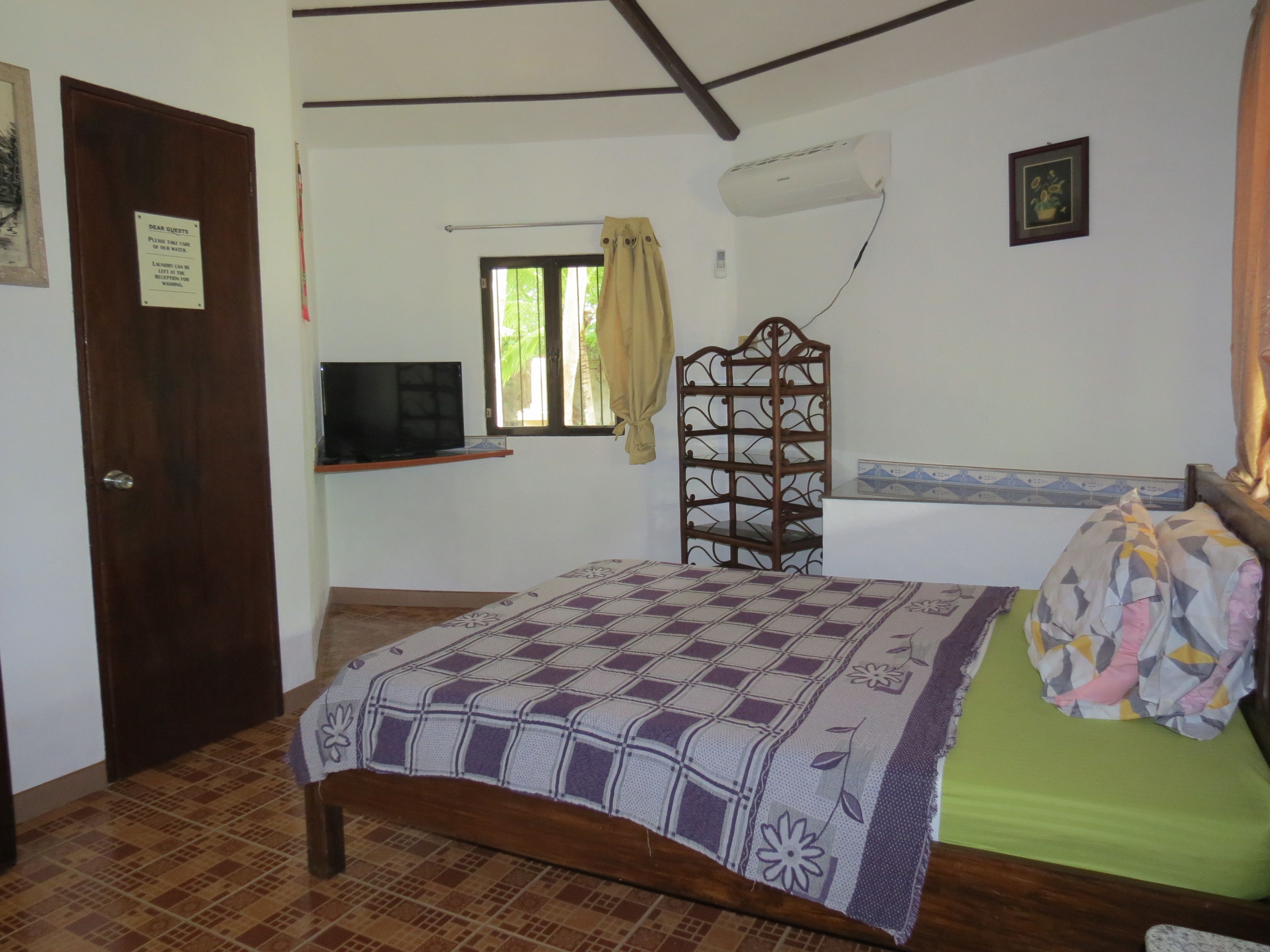 Un dormitorio con cama, televisión sobre una estantería, ventana con cortinas y equipo de aire acondicionado.