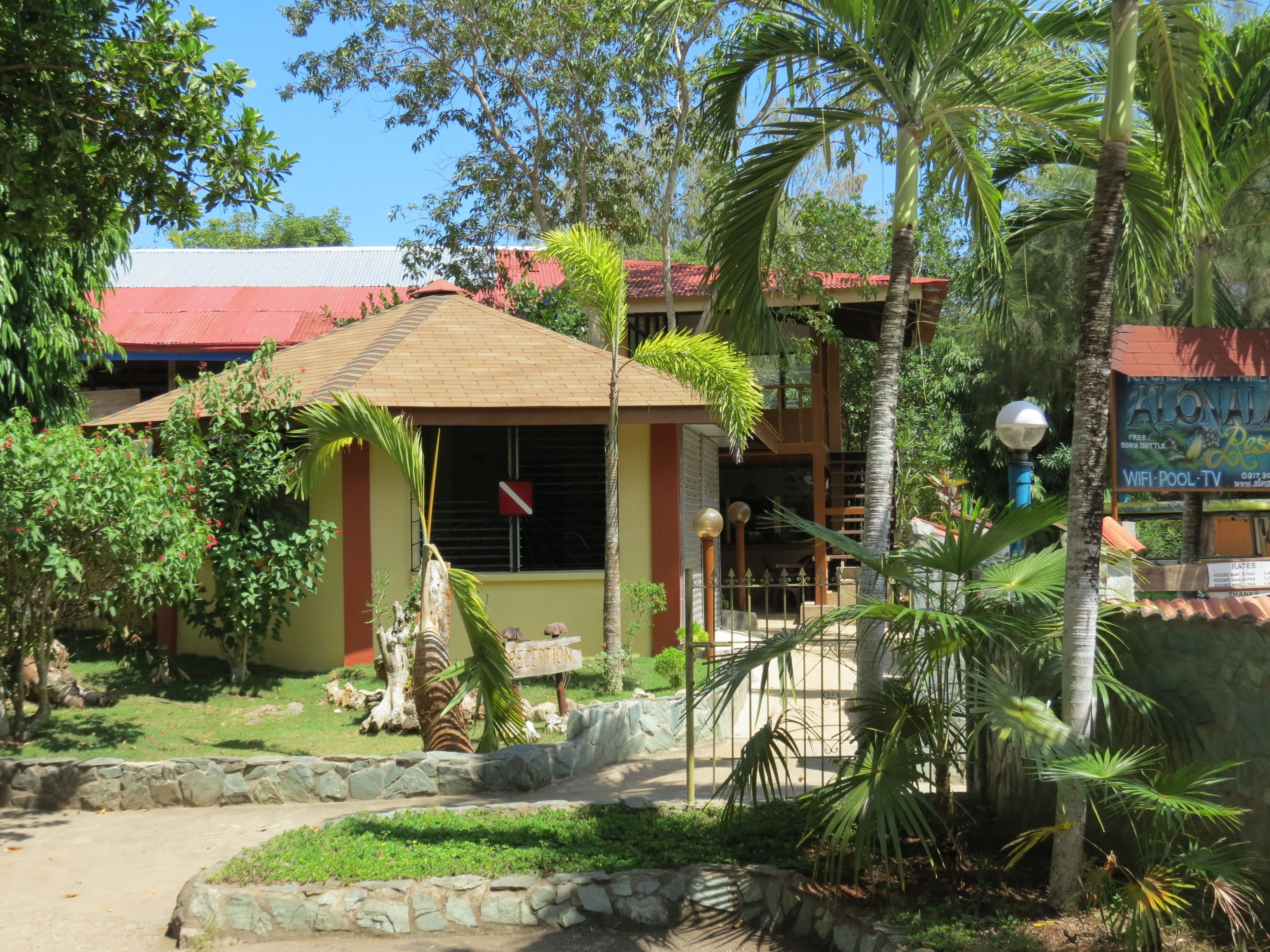 Un edificio de dos pisos con techo rojo, rodeado de abundante vegetación y palmeras.