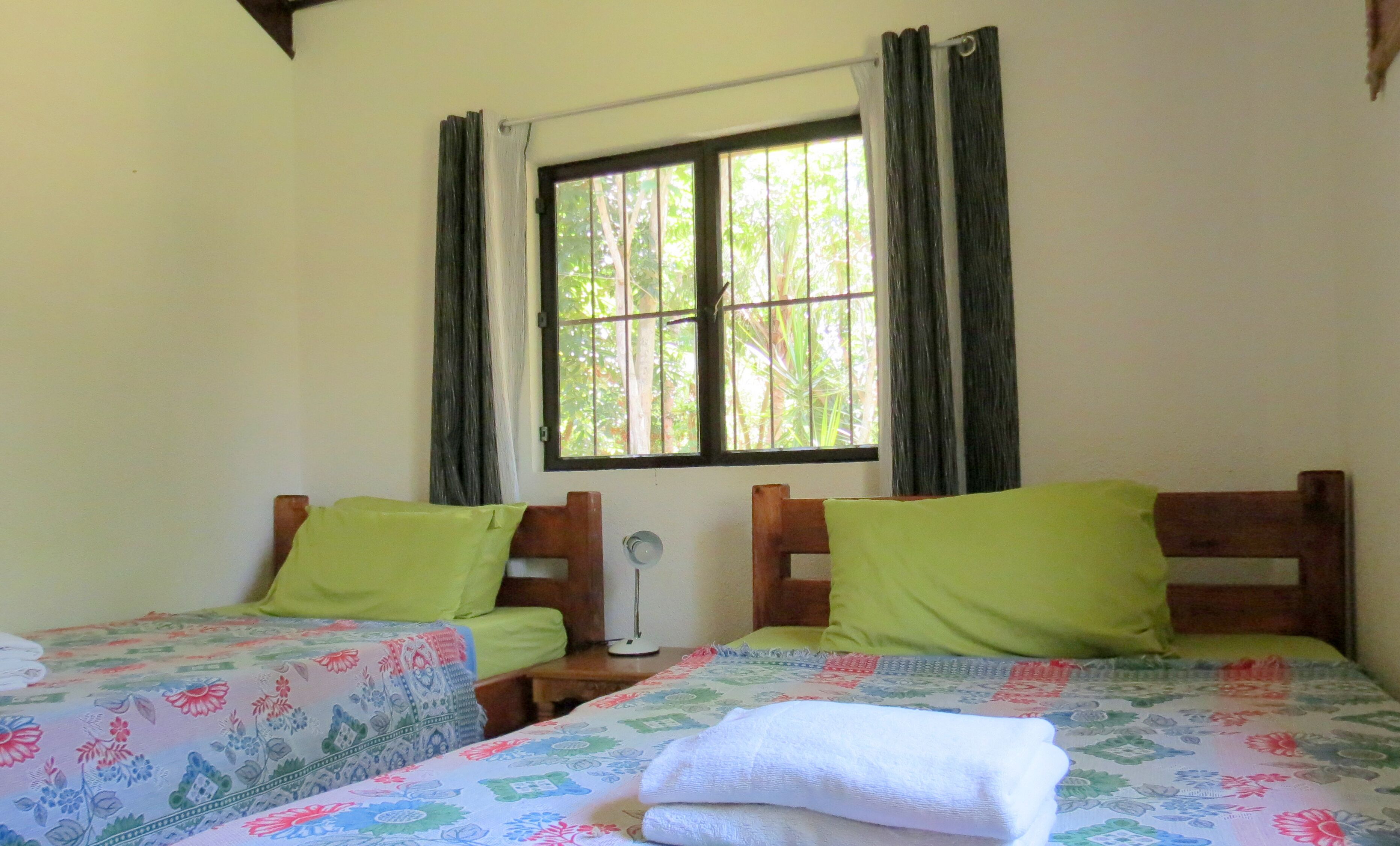 A room with two single beds, each with a floral-patterned bedspread and a green pillow. There is a window with curtains, a bedside table with a lamp, and a wall-mounted radiator.