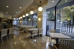 Um restaurante espaçoso com mesas e cadeiras de madeira, amplas janelas com vista para árvores e iluminação pendente.