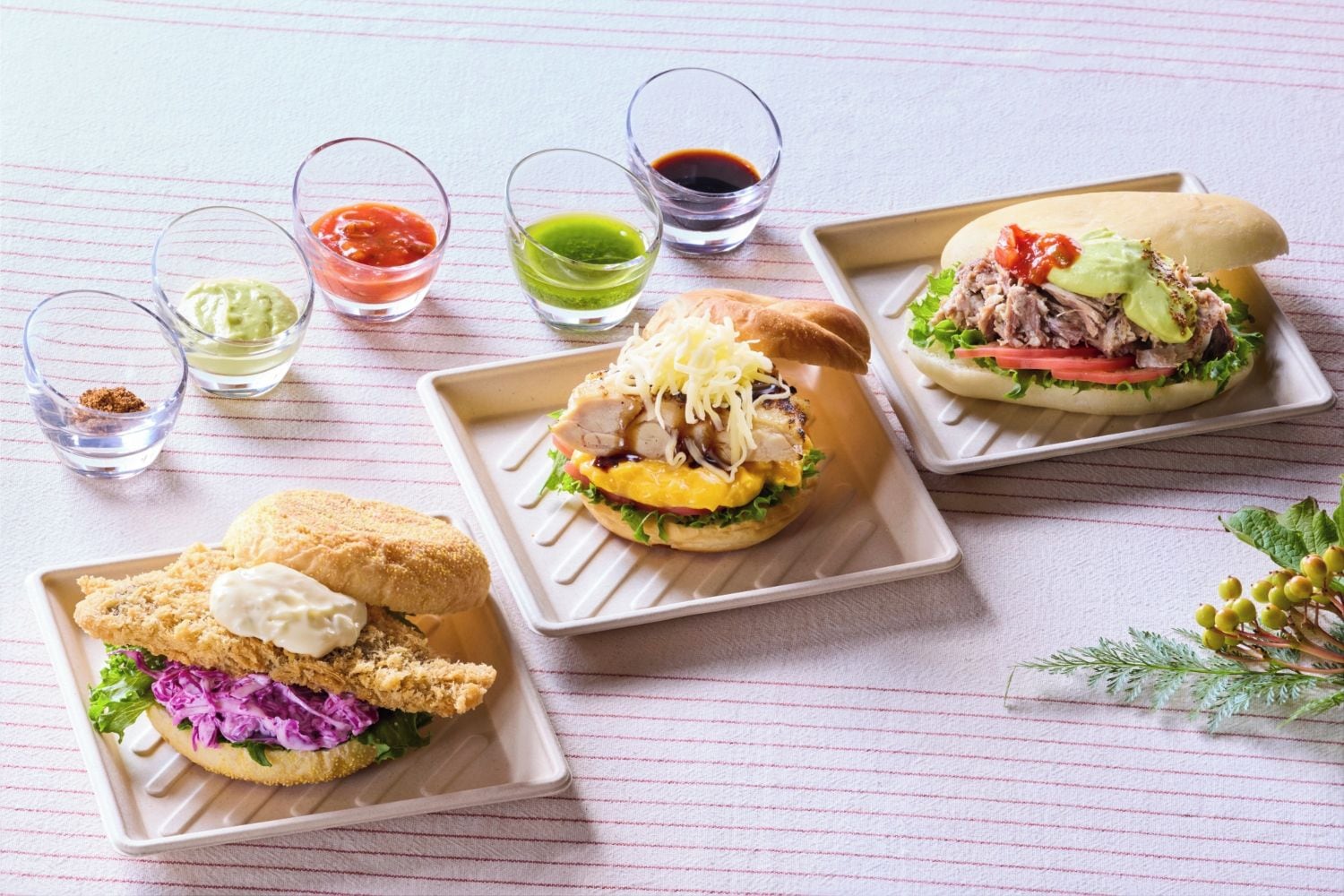 Three square plates with sandwiches, each accompanied by a small glass of sauce.