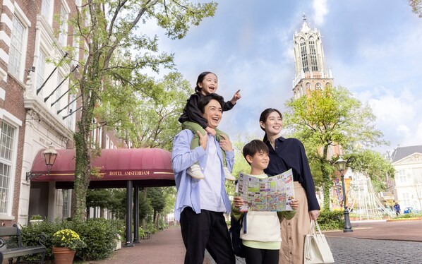 A family with a child on a man's shoulders, standing in front of a building with a red awning labeled "Hotel Amsterdam."