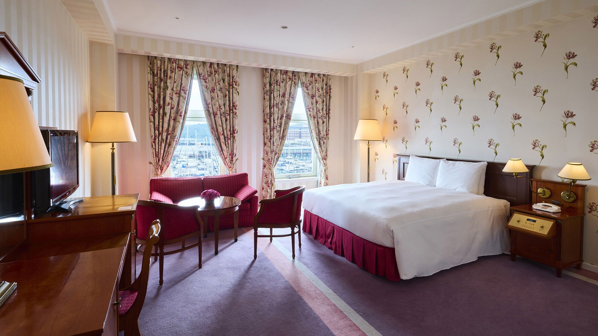 A hotel room with a bed, two bedside tables with lamps, a desk with a television, a chair, and a sofa.