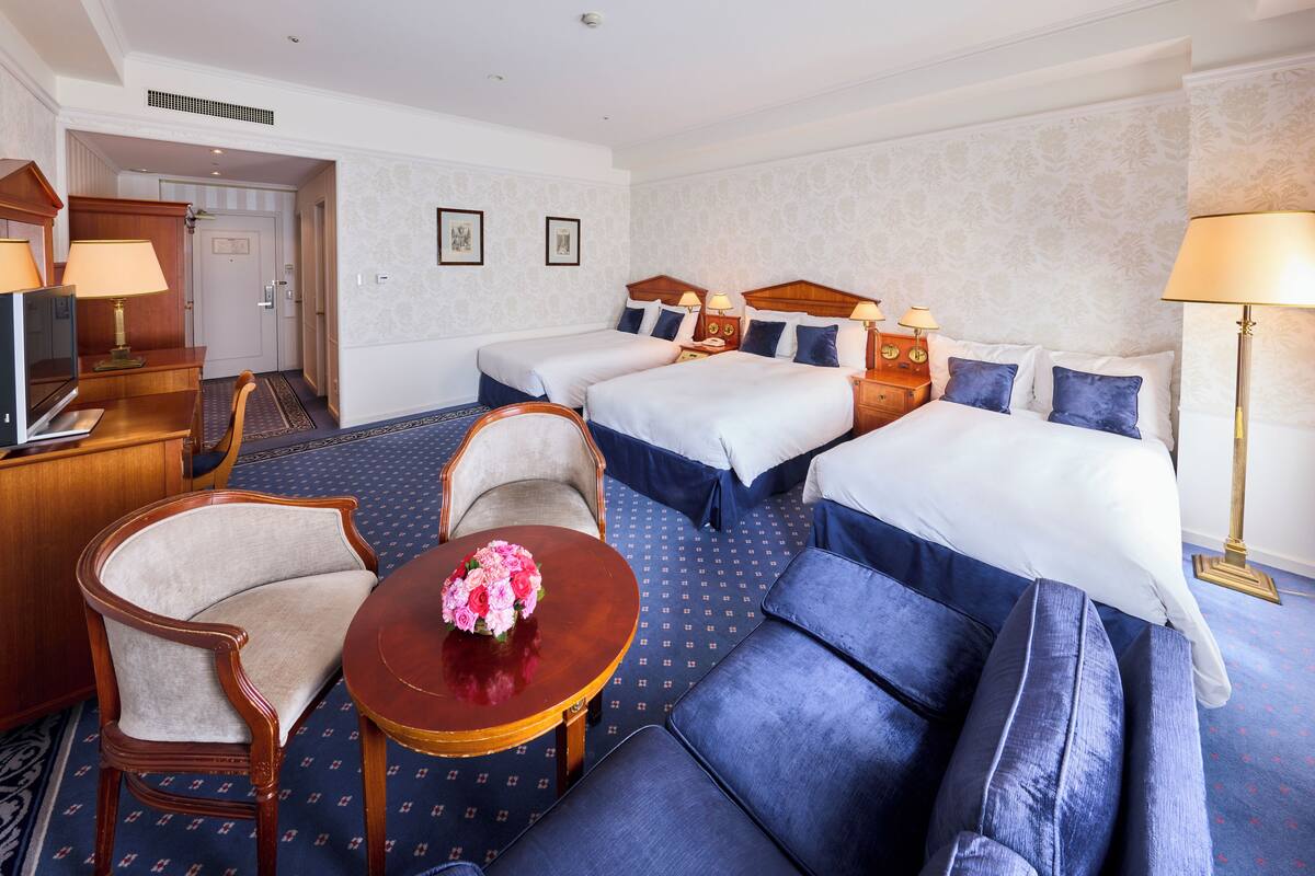 Une chambre d’hôtel avec deux lits, un canapé, un fauteuil, une petite table avec un vase et un bureau avec une lampe.