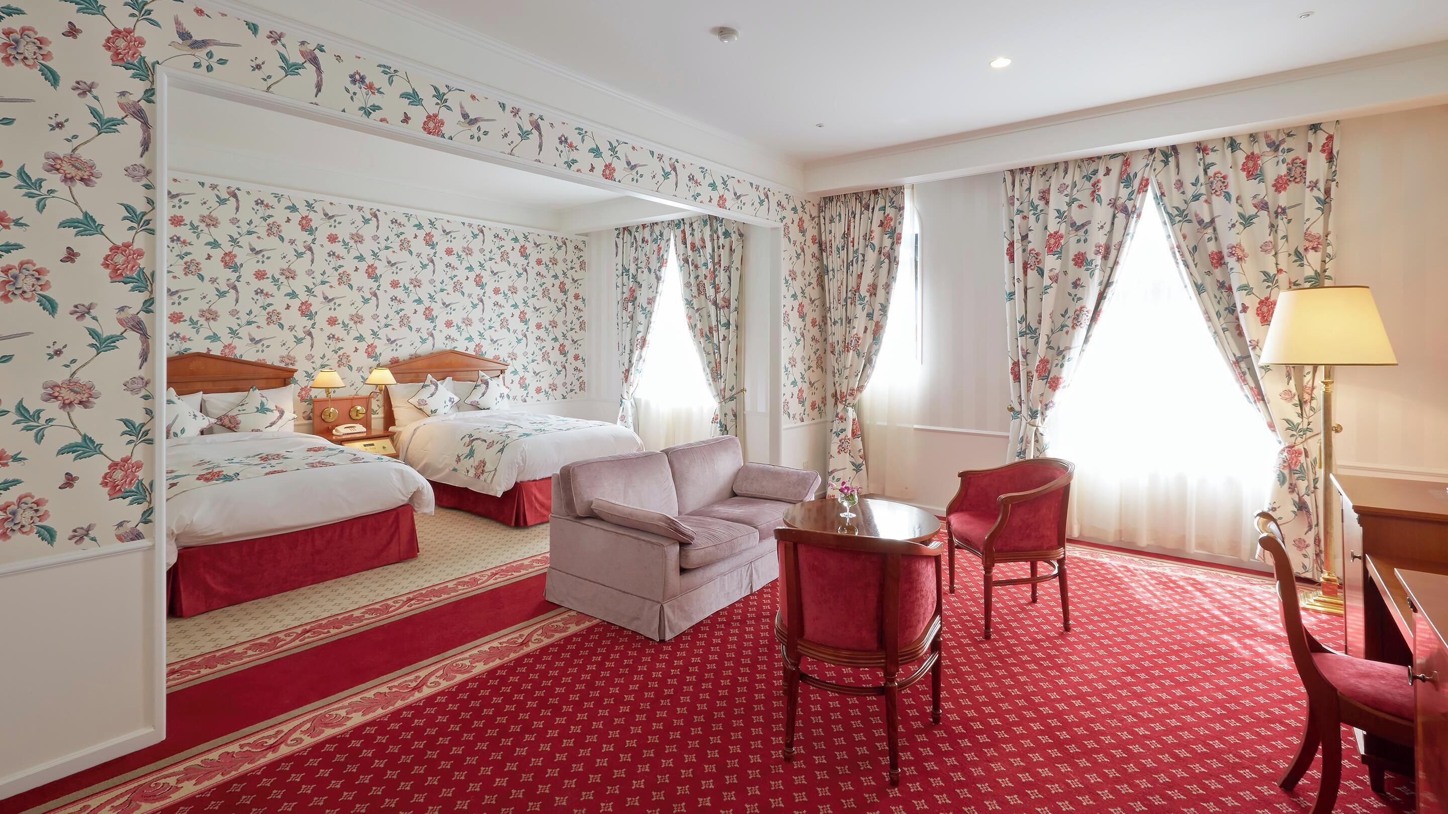 A hotel room with floral wallpaper, two beds, a sofa, a chair, a table, and a lamp.