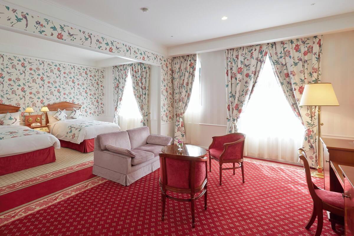 Une chambre d’hôtel avec un papier peint à motifs floraux, deux lits, un canapé, une chaise, une table et une lampe.