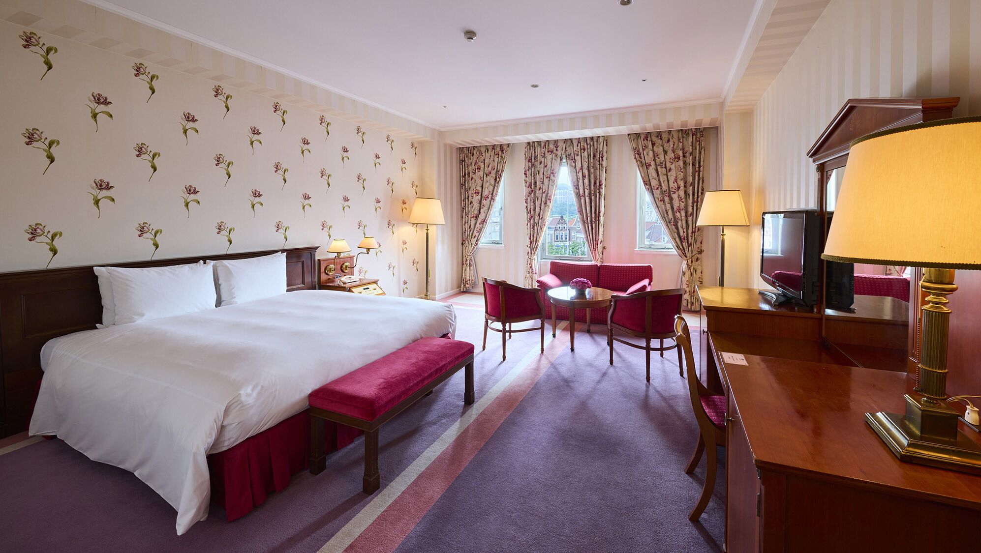 A hotel room with a large bed, a desk with a lamp, a chair, a sofa, and a television.