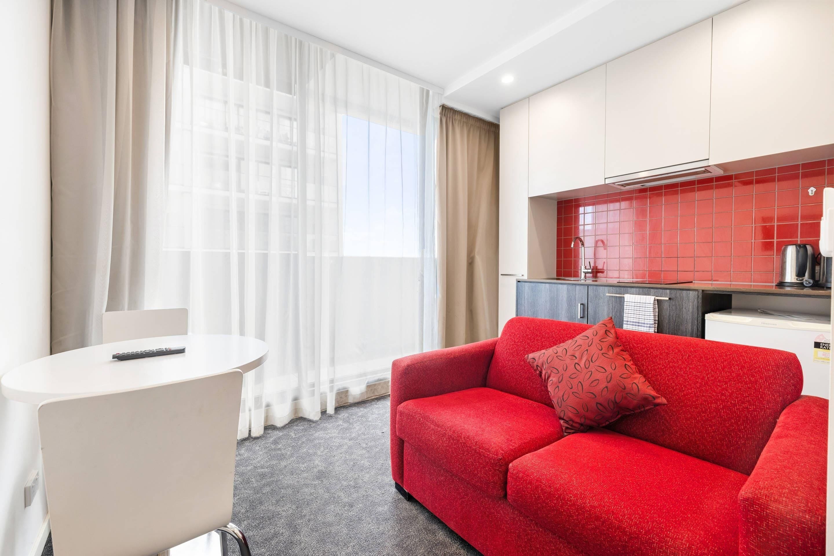 Une chambre d’hôtel avec un canapé rouge, une table blanche et une kitchenette carrelée de carreaux rouges.