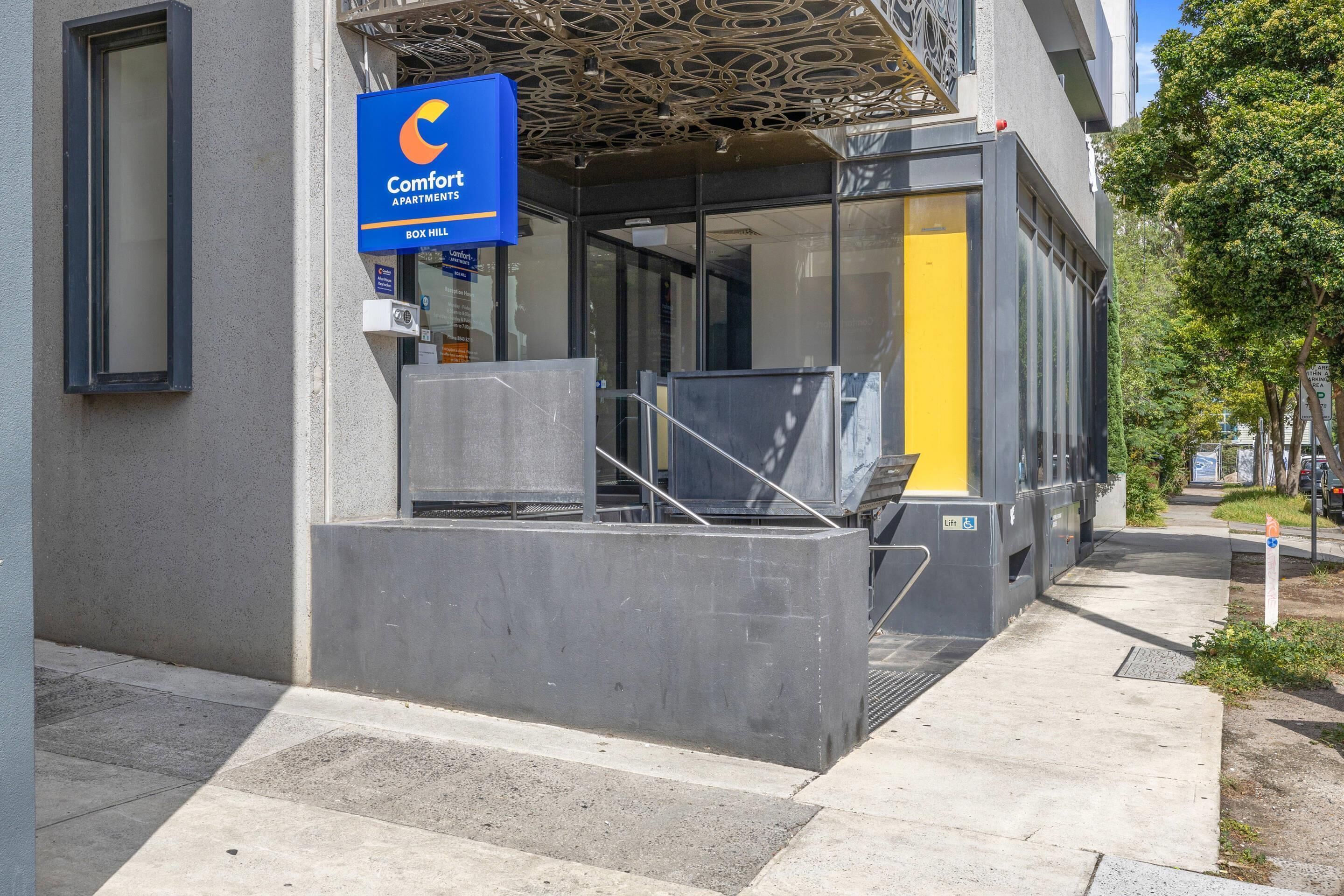 A modern building entrance with a blue sign for Comfort Apartments, a glass door, and a concrete ramp.