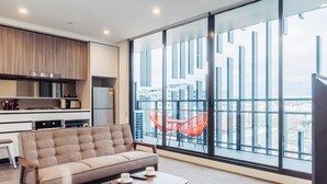 A modern living room with a sofa, a small table, and a large window with a view of buildings. - Comfort Apartments Box Hill (Box Hill)