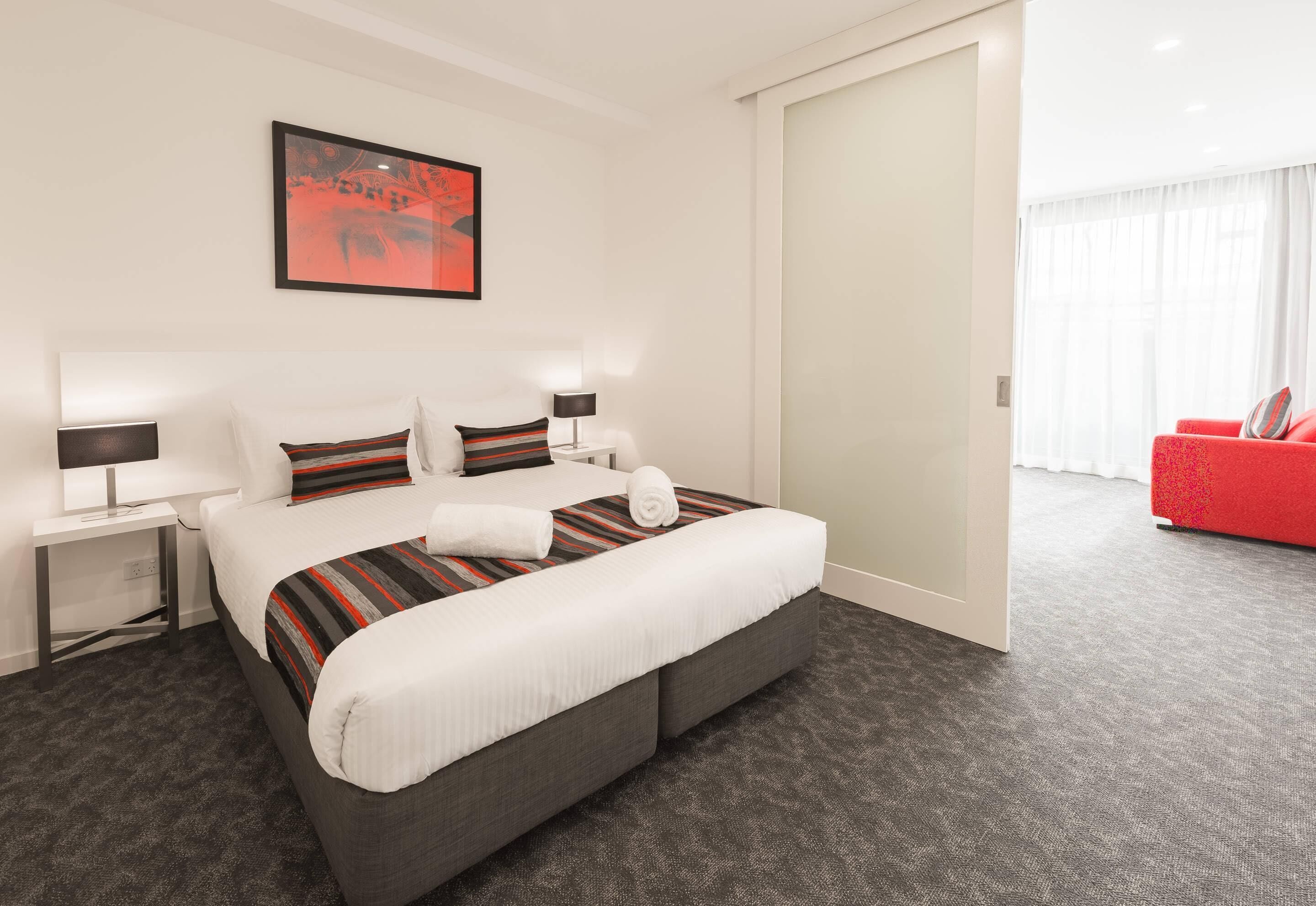 A hotel room with a bed, bedside tables, a red armchair, and a large window.
