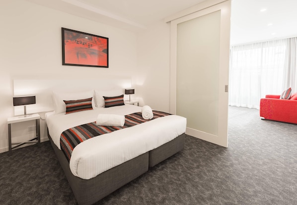 Una habitación de hotel con una cama, mesitas de noche, un sillón rojo y un ventanal grande.
