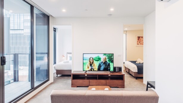 A modern living room with a large flat-screen TV, a sofa, a coffee table, and a sliding glass door leading to a balcony. - Comfort Apartments Box Hill (Box Hill)