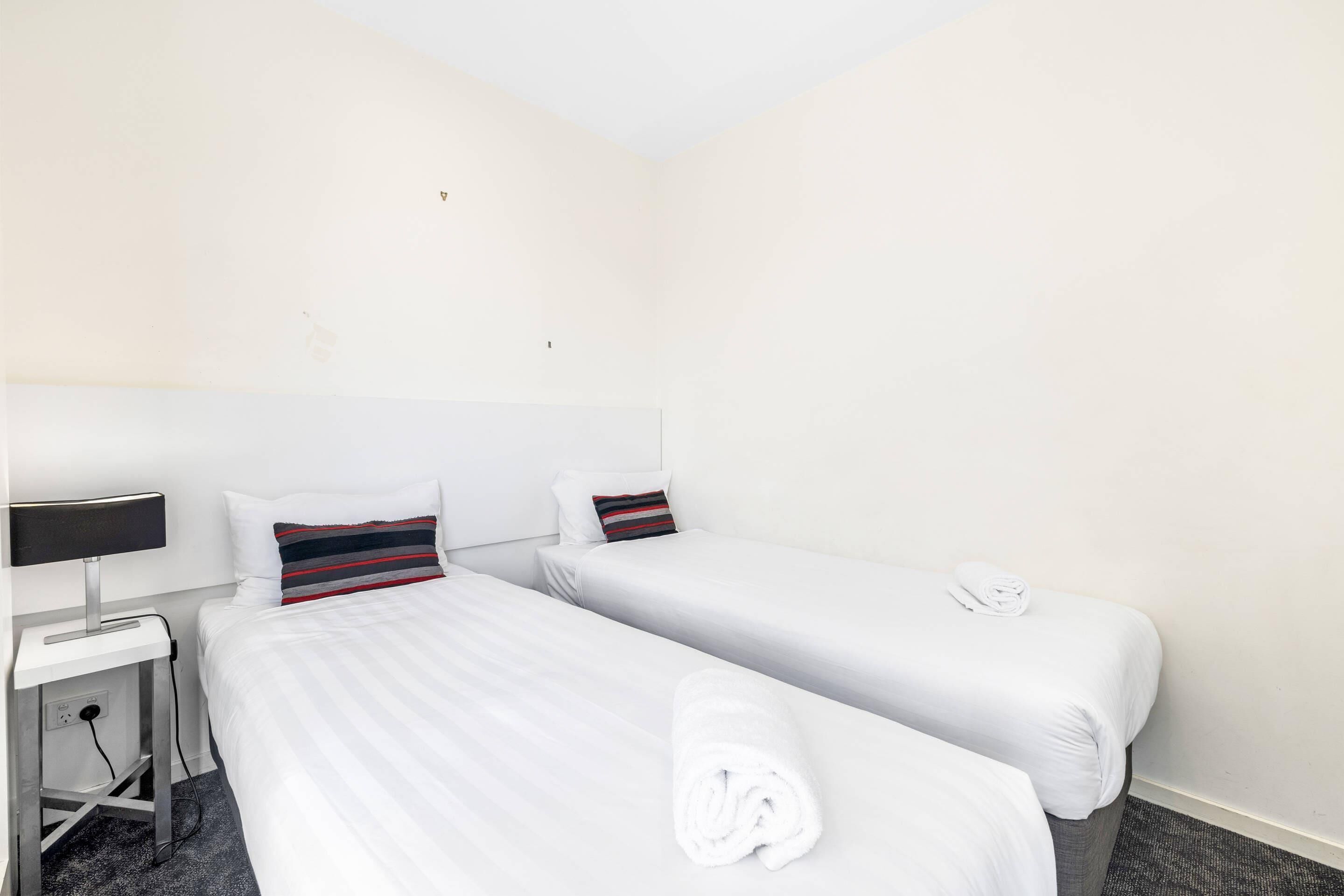 A hotel room with two single beds, a desk with a lamp, and a rolled towel on the floor.