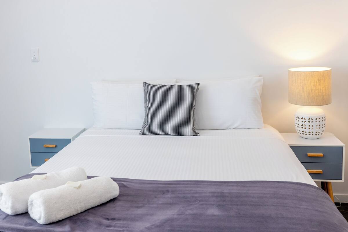 Un lit bien fait, avec une literie blanche et un oreiller à rayures, une lampe de chevet, un petit tiroir et deux serviettes posées sur le lit.