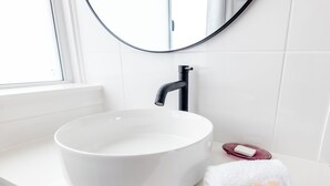 Une salle de bain avec un lavabo blanc, un miroir rond et une fenêtre.