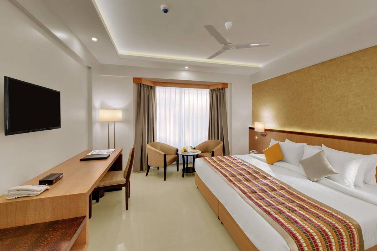 A hotel room with a bed, desk, chairs, a TV, and a ceiling fan.